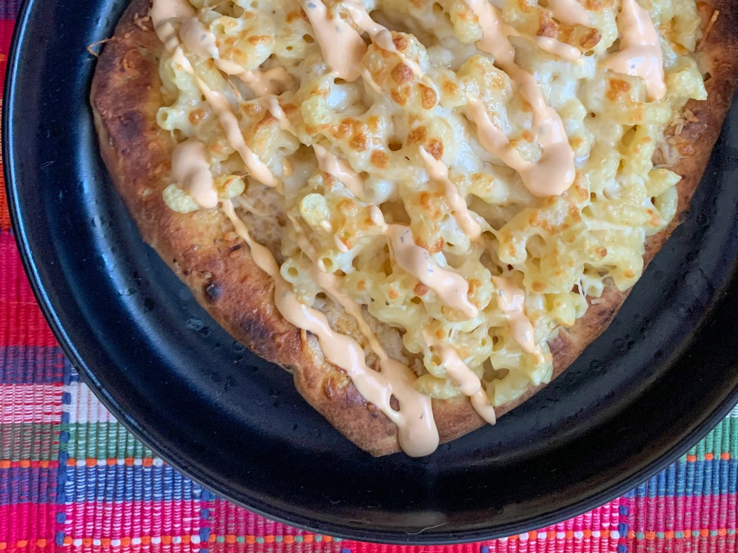 Elbow mac and cheese topped with an orange sauce on a naan crust on a black bowl with a pink plaid background