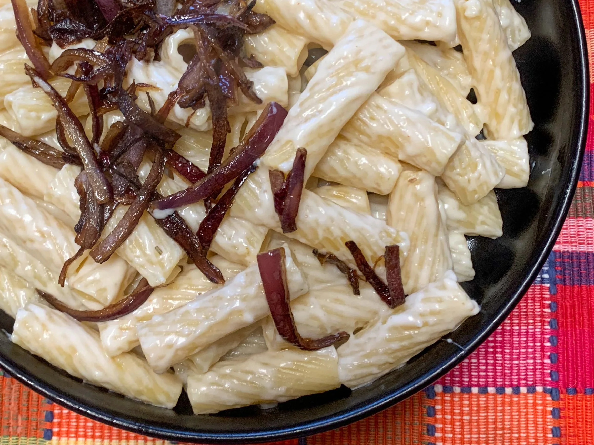 Tube shaped pasta in a white sauce topped with purple onions in a black bowl on a pink plaid background to make hatch chile mac and cheese