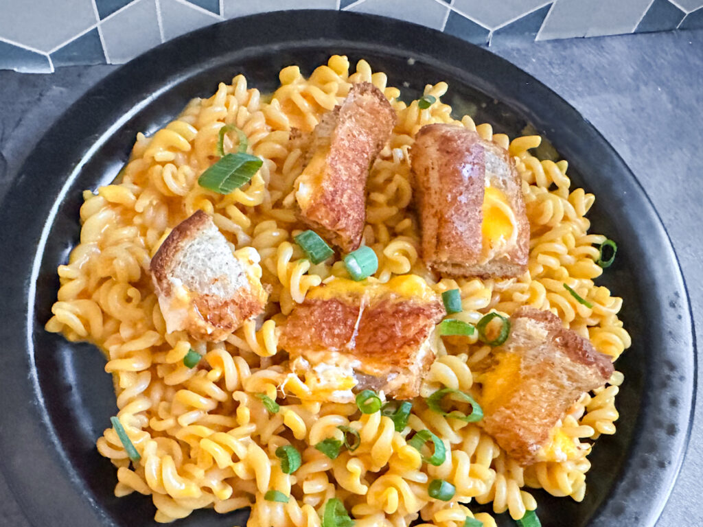 Bowl of Thrilled Cheese mac and cheese topped with chopped green onions and crispy grilled cheese croutons, served on a black plate with a geometric tile background