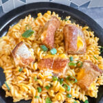 Bowl of Thrilled Cheese mac and cheese topped with chopped green onions and crispy grilled cheese croutons, served on a black plate with a geometric tile background
