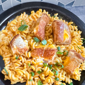 Bowl of Thrilled Cheese mac and cheese topped with chopped green onions and crispy grilled cheese croutons, served on a black plate with a geometric tile background