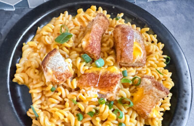 Bowl of Thrilled Cheese mac and cheese topped with chopped green onions and crispy grilled cheese croutons, served on a black plate with a geometric tile background