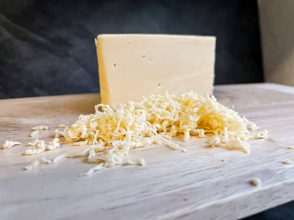 A block of gouda cheese and a small pile of shredded gouda on a light wooden cutting board, with a dark tiled background.