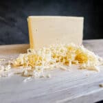 A block of gouda cheese and a small pile of shredded gouda on a light wooden cutting board, with a dark tiled background.