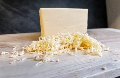 A block of gouda cheese and a small pile of shredded gouda on a light wooden cutting board, with a dark tiled background.